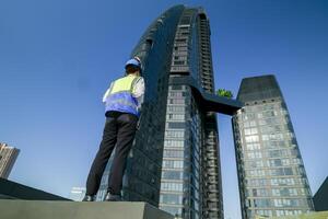 Engineer with tablet on rooftop assessing HVAC system against city skyline, safety equipment and hard hat on. photo