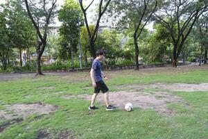 asiático hombre son corriendo y pateando pelota para jugando un fútbol a al aire libre parque. foto