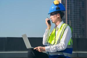 Focused engineer in hard hat and safety vest using walkie-talkie and laptop on construction site with urban backdrop. photo
