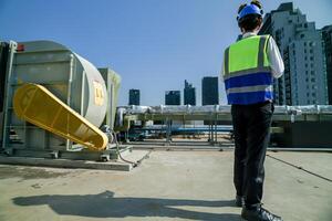 Skilled engineer operating tablet to manage HVAC machinery on top of building, urban architecture in distance. photo