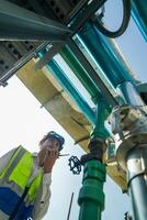 Industrial engineer adjusting pipeline valve with radio communication on construction site. Construction worker using walkie-talkie to coordinate machinery adjustments on a sunny day. photo