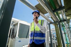 Industrial engineer adjusting pipeline valve with radio communication on construction site. Engineer on urban construction site using radio to coordinate with team on a sunny industrial rooftop. photo