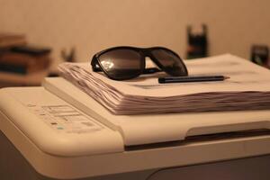 Sunglasses with pen lie on a stack of paper on the printer photo