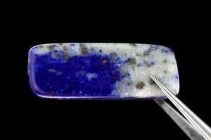 Lapis lazuli jewel stone texture in tweezers on black background. Macro closeup. photo