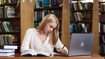 atraente multiétnico aluna fazer uma haste classe dever de casa dentro uma livro didático, enquanto pesquisando questionário respostas em uma computador portátil computador. jovem Loiras fêmea estudando dentro uma público biblioteca com clássico interior video