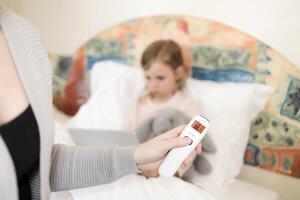 mother measuring temperature of her daughter. child got high temperature. Kid sitting in bed with tablet and furry toy and no face woman holding infrared thermometer which indicates 38,2 C photo