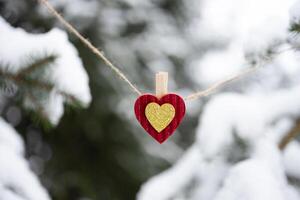 Red Christmas ornament in shape of heart hanging on jute twine string on snow-covered fir tree background outdoors. Concept of love, family, support photo