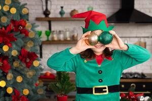 Child holding up two Christmas ornaments in front of her eyes. Little girl dressed up as elf. Blurred Christmas tree with bokeh lights and other holiday decorations on background photo