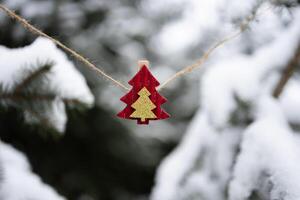 Red Christmas ornament in shape of fir tree hanging on jute twine string on snow-covered Christmas trees background outdoors. Selective focus, close-up photo