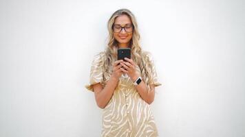 Happy woman in trendy eyeglasses using smartphone in front of white wall video