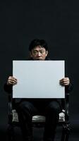 A Chinese man sits in a chair, solemnly holding a blank white sign photo
