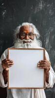 An elderly Indian man smiling, holding a blank white board in front of a blackboard photo