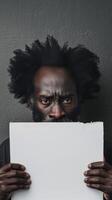 A mature man with dreadlocks and a black afro looks pensive while holding up a blank white paper photo