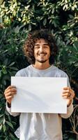 A man gleefully holds a white sign in front of a lush bush photo