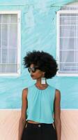A stylish black woman stands in front of a bright turquoise wall, wearing sunglasses and a turquoise top photo