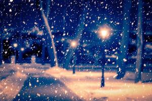 Blured photo of a winter night park with lanterns, pavement and trees covered with snow in snowfall.