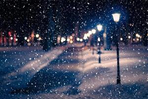 Blured photo of a winter night park with lanterns, pavement and trees covered with snow in snowfall.
