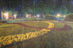 Defocused night park with lanterns and flowers behind rainy, wet glass at night. Concept of rainy autumn weather. photo