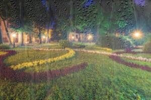 Defocused night park with lanterns and flowers behind rainy, wet glass at night. Concept of rainy autumn weather. photo