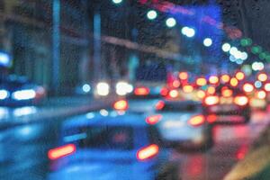 Blurry moving cars with headlights and lanterns reflecting on the wet asphalt in the night city behind rain-covered window. photo