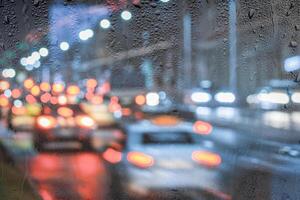 Blurry moving cars with headlights and lanterns reflecting on the wet asphalt in the night city behind rain-covered window. photo