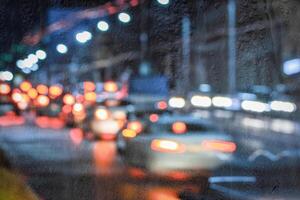 Blurry moving cars with headlights and lanterns reflecting on the wet asphalt in the night city behind rain-covered window. photo