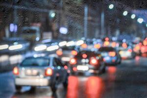 Blurry moving cars with headlights and lanterns reflecting on the wet asphalt in the night city behind rain-covered window. photo