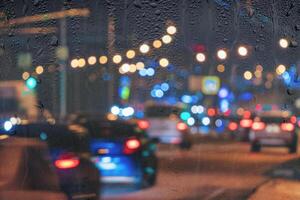 Blurry moving cars with headlights and lanterns reflecting on the wet asphalt in the night city behind rain-covered window. photo