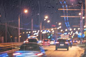 Blurry moving cars with headlights and lanterns reflecting on the wet asphalt in the night city behind rain-covered window. photo