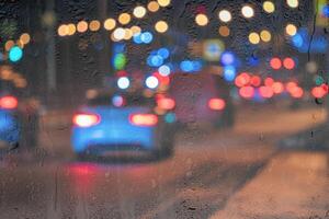 Blurry moving cars with headlights and lanterns reflecting on the wet asphalt in the night city behind rain-covered window. photo