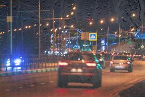 Blurry moving cars with headlights and lanterns reflecting on the wet asphalt in the night city behind rain-covered window. photo
