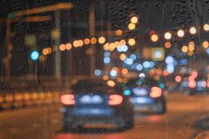 Blurry moving cars with headlights and lanterns reflecting on the wet asphalt in the night city behind rain-covered window. photo