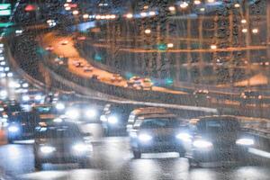 Blurry moving cars with headlights and lanterns reflecting on the wet asphalt in the night city behind rain-covered window. photo