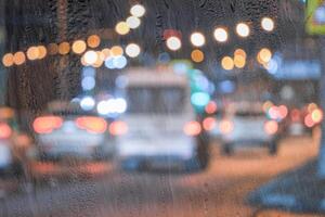 Blurry moving cars with headlights and lanterns reflecting on the wet asphalt in the night city behind rain-covered window. photo