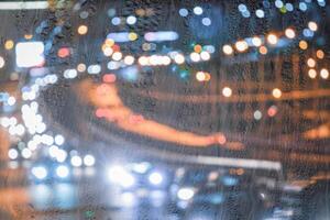 Blurry moving cars with headlights and lanterns reflecting on the wet asphalt in the night city behind rain-covered window. photo