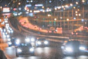 Blurry moving cars with headlights and lanterns reflecting on the wet asphalt in the night city behind rain-covered window. photo