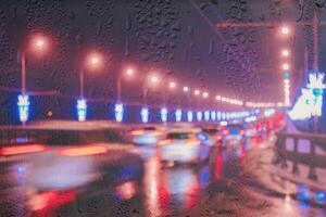 Blurry lights of moving cars and lanterns reflecting on the wet asphalt in the night city behind rain-covered window. photo