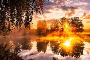 Misty sunrise on the pond in the autumn morning. Birch trees with rays of the sun cutting through the branches, reflected in the water. photo