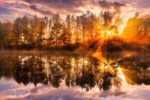 Misty sunrise on the pond in the autumn morning. Trees with rays of the sun cutting through the branches, reflected in the water. photo