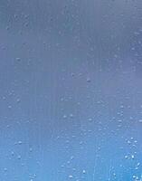 Rain drops on window glasses surface with blue sky and clouds background. Raindrops on a glass. The concept of changing from rainy to sunny weather. photo