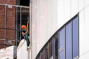 Russia, Kaluga - SEPTEMBER 16, 2020 An industrial climber hanging from a rope with a carabiner and securing the white cladding panels of a building at a construction site. photo