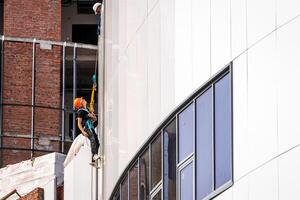 Russia, Kaluga - SEPTEMBER 16, 2020 An industrial climber hanging from a rope with a carabiner and securing the white cladding panels of a building at a construction site. photo
