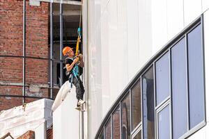Russia, Kaluga - SEPTEMBER 16, 2020 An industrial climber hanging from a rope with a carabiner and securing the white cladding panels of a building at a construction site. photo