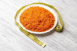 Grains of red lentils and a measuring tape in a plate on a white wooden table. photo