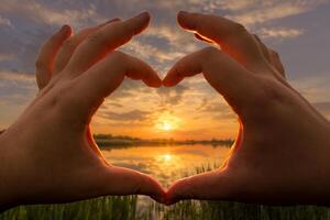 Hands in the shape of heart against the sunset over the lake or river in the summer with a cloudy sky background. Landscape. photo
