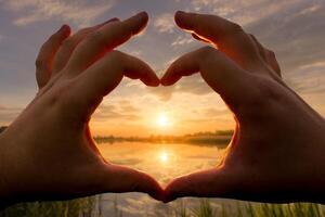 Hands in the shape of heart against the sunset over the lake or river in the summer with a cloudy sky background. Landscape. photo