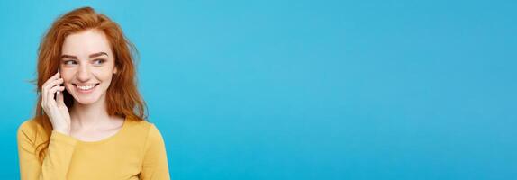 Lifestyle and Technology concept - Portrait of cheerful happy ginger red hair girl with joyful and exciting talking with friend by mobile phone. Isolated on Blue Pastel Background. Copy space. photo