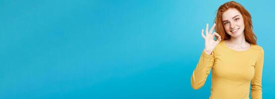 Fun and People Concept - Headshot Portrait of charming ginger red hair girl with freckles smiling and making ok sign with finger. Pastel blue background. Copy Space. photo