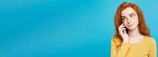 Lifestyle and Technology concept - Portrait of ginger red hair girl with shocking and stressful expression while talking with friend by mobile phone. Isolated on Blue Pastel Background. Copy space. photo