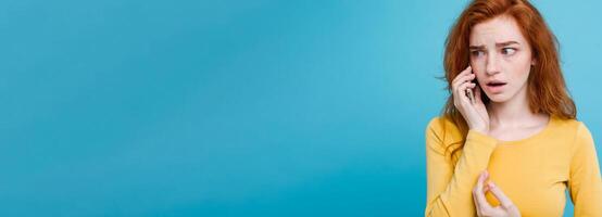 Lifestyle concept - Portrait of ginger red hair girl with shocking and stressful expression while talking with friend by mobile phone. Isolated on Blue Pastel Background. Copy space photo